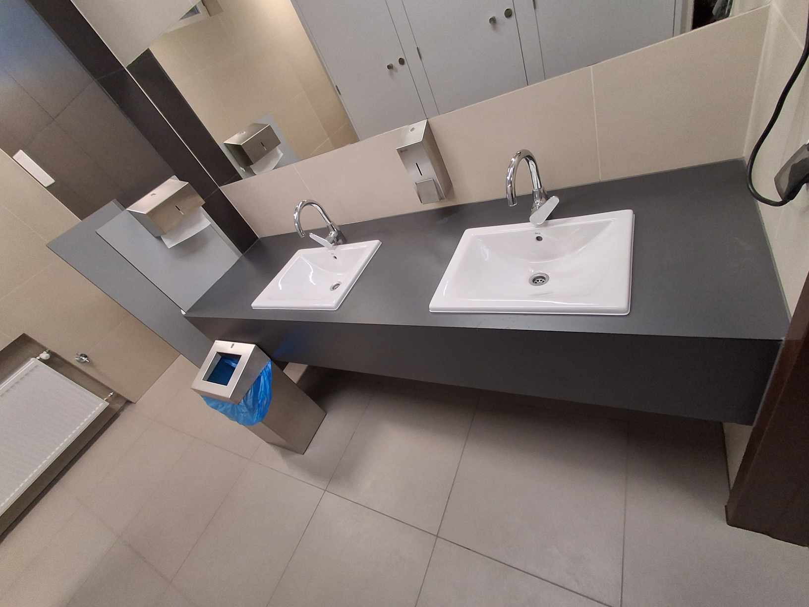 Dwie białe umywalki nablatowe z chromowanymi bateriami na szarym blacie w publicznej toalecie, z dozownikami mydła i ręczników papierowych na ścianie.
