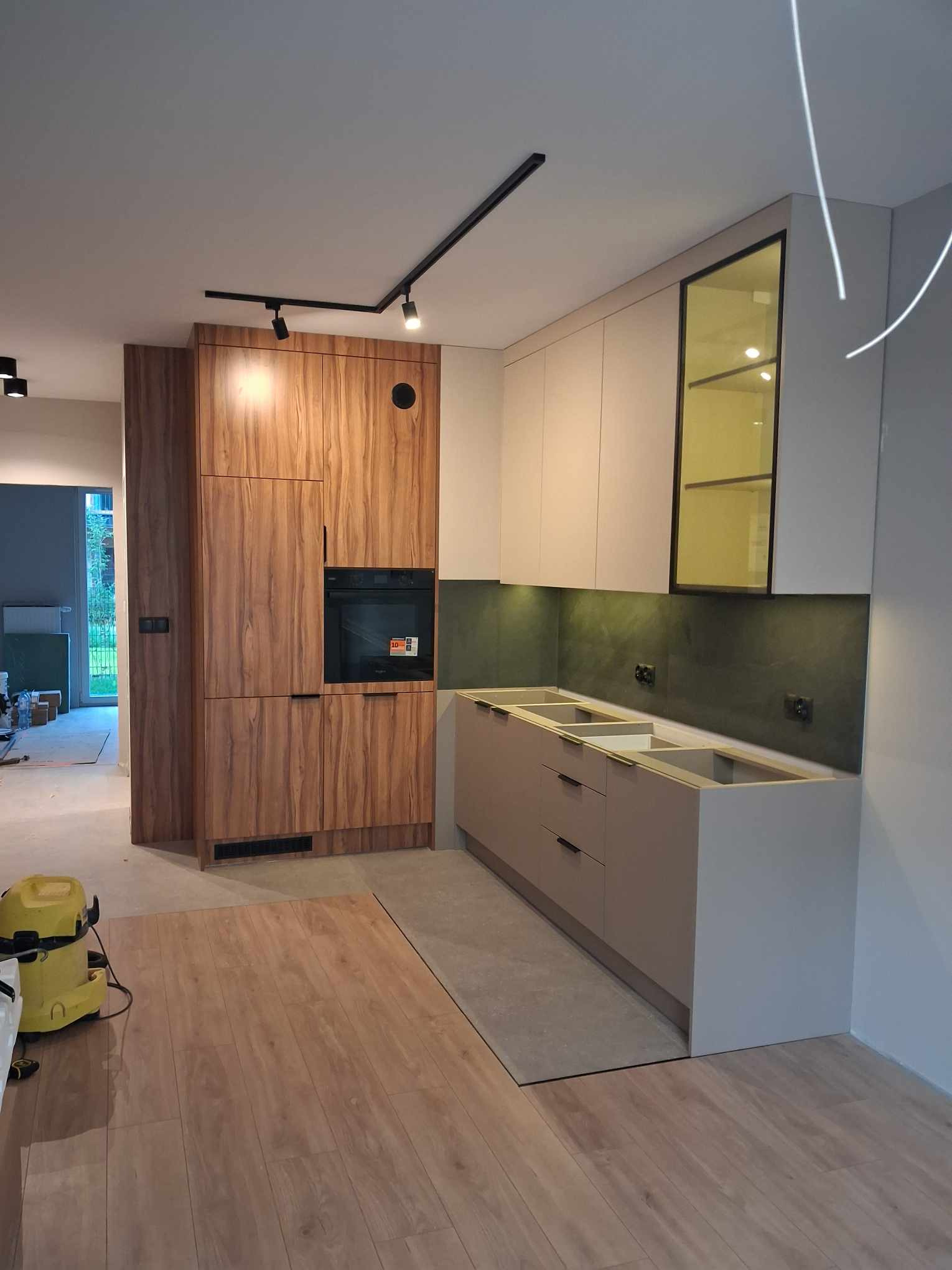Montaż kuchni na wymiar w trakcie: drewnopodobna zabudowa z piekarnikiem, szare szafki dolne bez blatów, zielony panel ścienny, czarne oświetlenie szynowe, żółty odkurzacz na podłodze.
