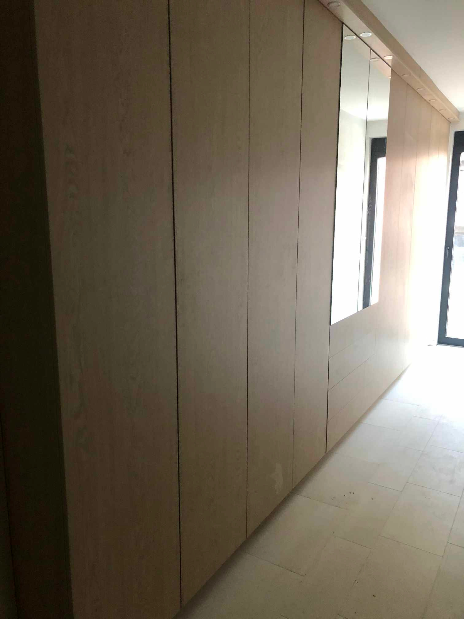 Długa, zabudowana szafa z jasnego drewna z wbudowanym lustrem, ciągnąca się wzdłuż korytarza z jasną podłogą i oknem w tle.