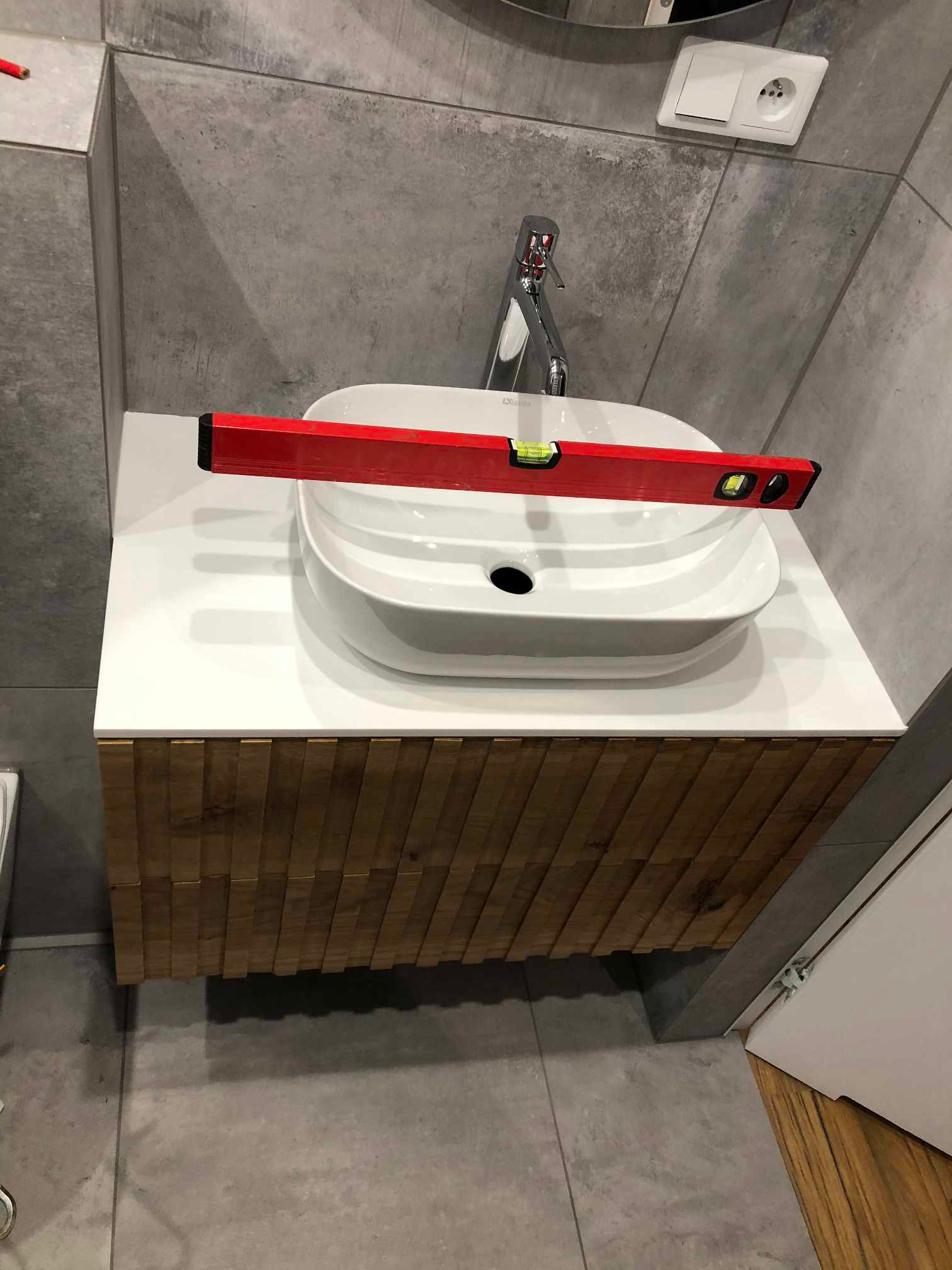 Biała, owalna umywalka nablatowa z baterią, ustawiona na szafce z drewnianymi lamelami; poziomica spoczywa na krawędzi umywalki, sprawdzając jej wypoziomowanie na blacie; ściana wykończona szarą...