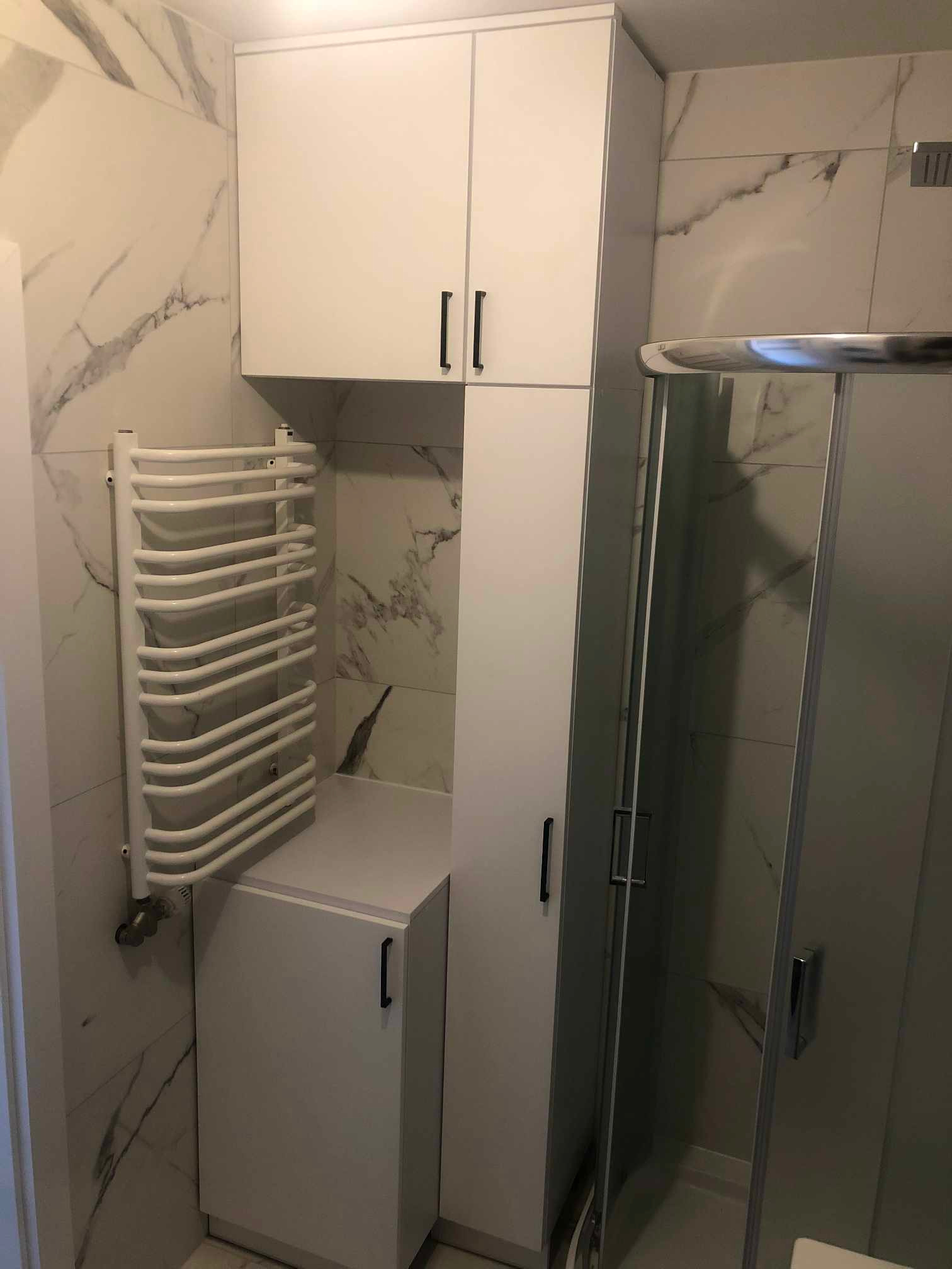 Biała zabudowa meblowa w łazience z marmurowymi płytkami, obejmująca szafki wiszące, słupek i szafkę pod umywalkę, obok grzejnik drabinkowy i kabina prysznicowa.