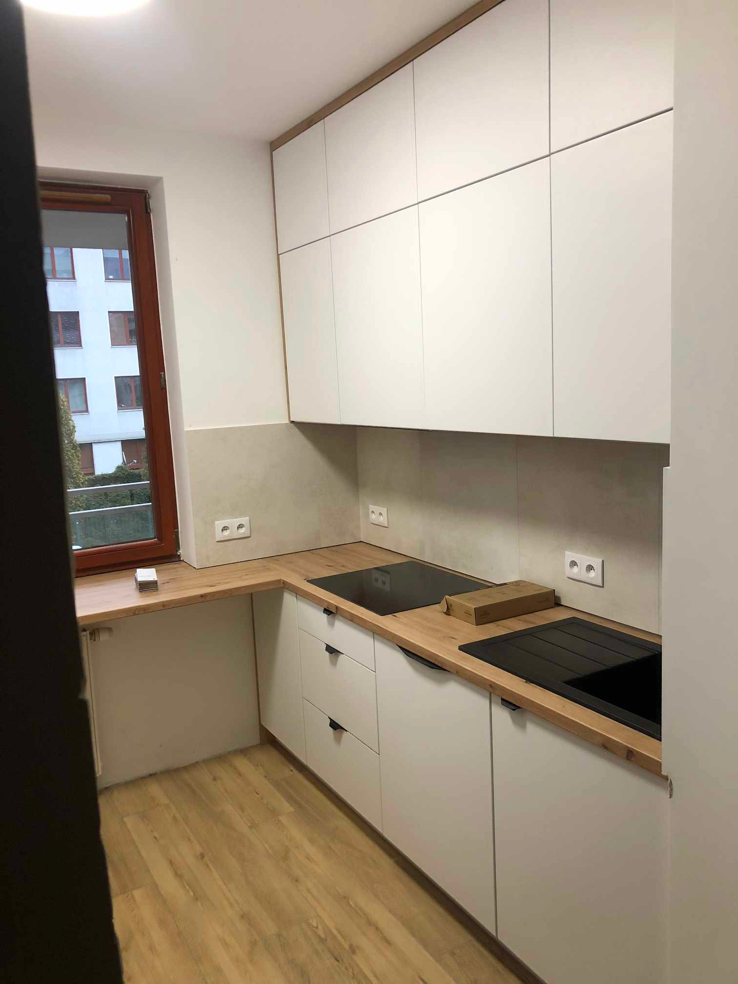 Nowoczesna kuchnia z białymi szafkami i drewnianym blatem, widok na okno z widokiem na budynek mieszkalny, płyta indukcyjna i zlew w kolorze czarnym.