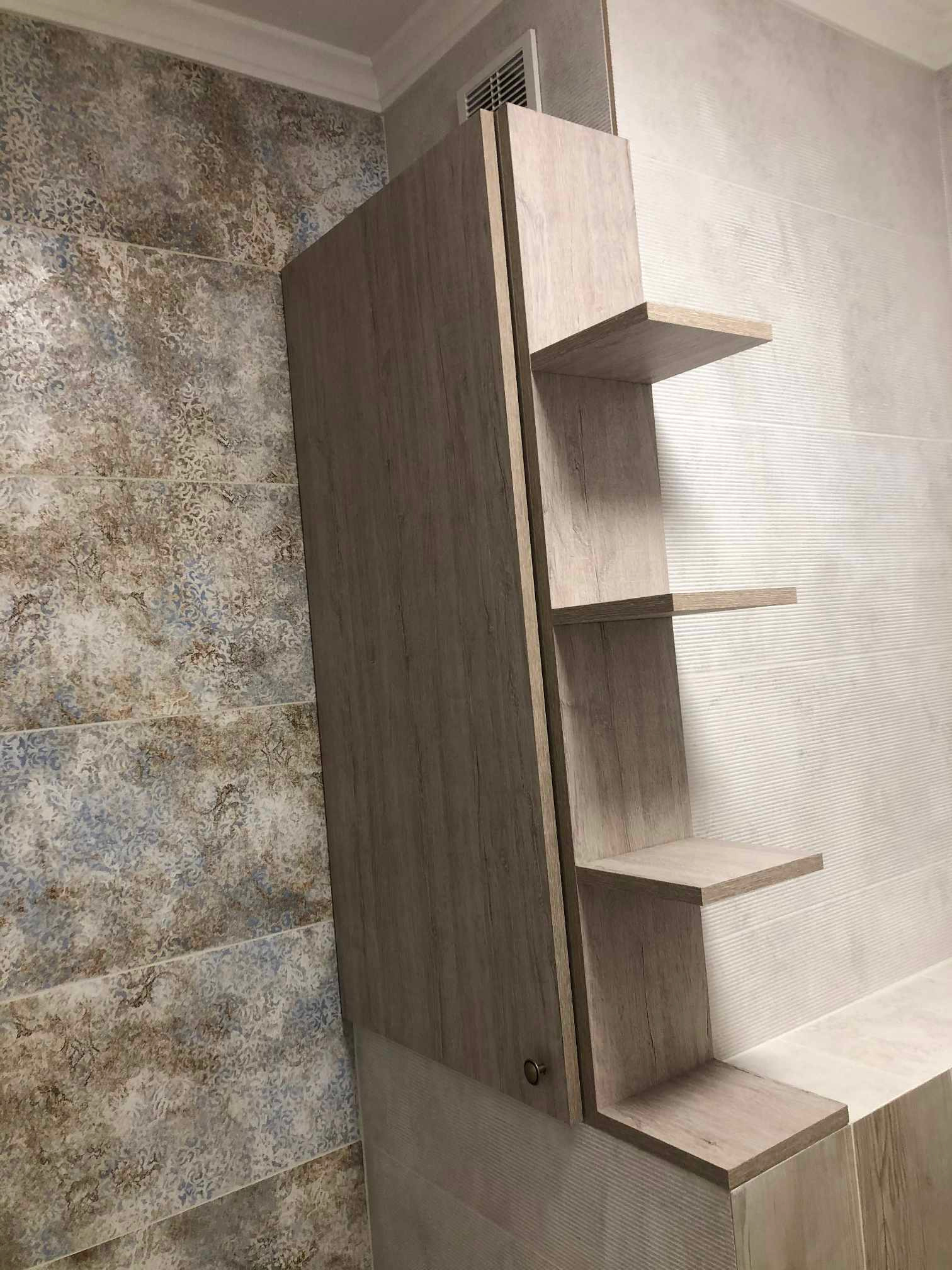 Jasna, drewnopodobna szafka łazienkowa z otwartymi półkami na tle płytek w dwóch wzorach: wzorzystych i z delikatnymi prążkami.