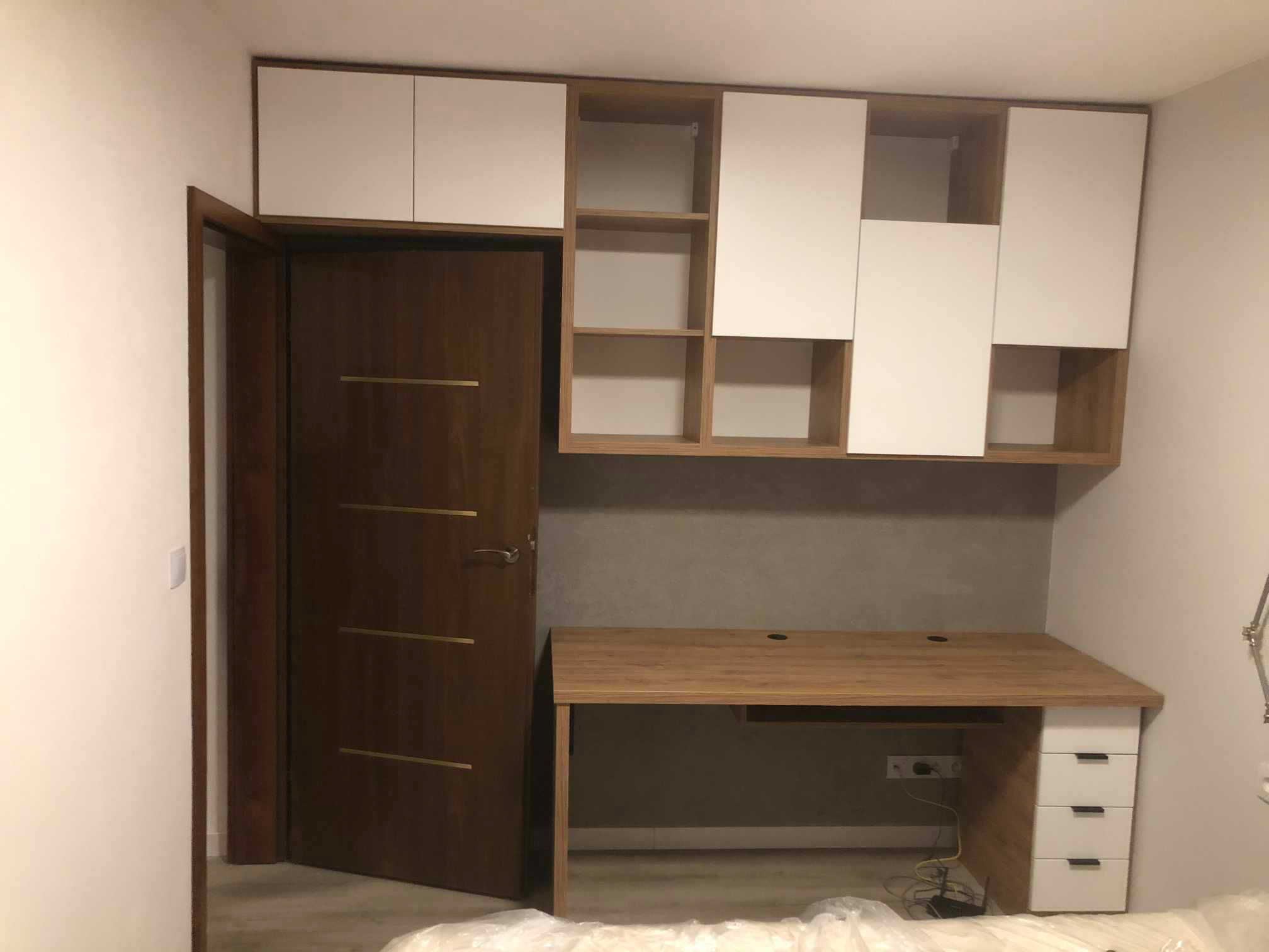 Biało-drewniana szafa wisząca z otwartymi półkami nad biurkiem z trzema szufladami, obok ciemnych drzwi z poziomymi, złotymi wstawkami.