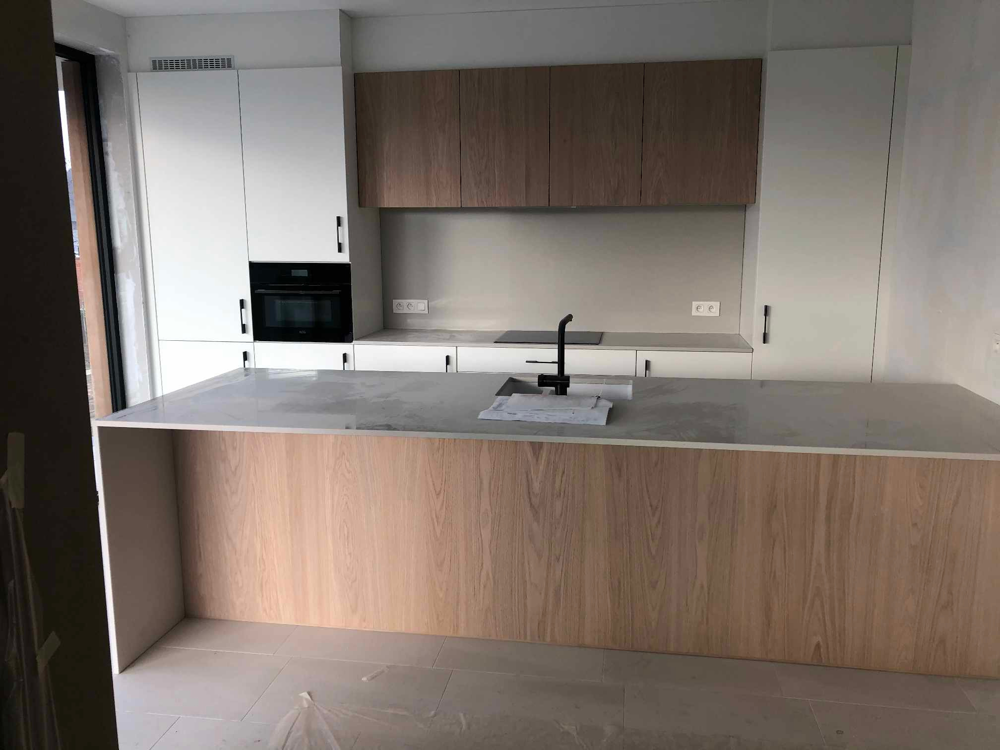 Nowoczesna kuchnia z wyspą, fronty szafek w kolorze dębu, białe szafki wiszące, czarny piekarnik i bateria.