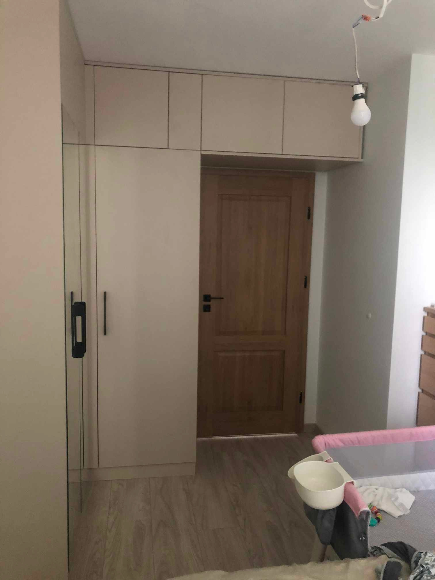 Jasna zabudowa szafy z górnymi szafkami nad drewnianymi drzwiami w pokoju z dziecięcym łóżeczkiem i wiszącą żarówką bez klosza.