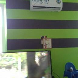 Syjmar - Klimatyzator ścienny zamontowany na zielono-szarej ścianie w pokoju, widoczny telewizor i krzesełko dziecięce.