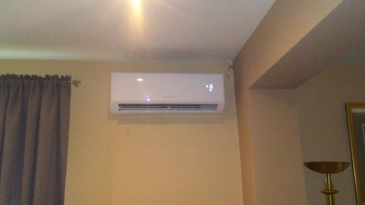 Biały klimatyzator ścienny zamontowany w pokoju z beżowymi ścianami, obok zasłoniętego okna i ozdobnej lampy. Na urządzeniu widoczny wyświetlacz z temperaturą.