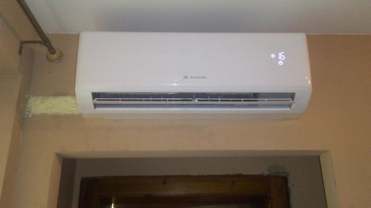 Biała jednostka klimatyzacji Alpicair zamontowana na ścianie nad futryną drzwi. Widoczne ślady pianki montażowej wokół rur.