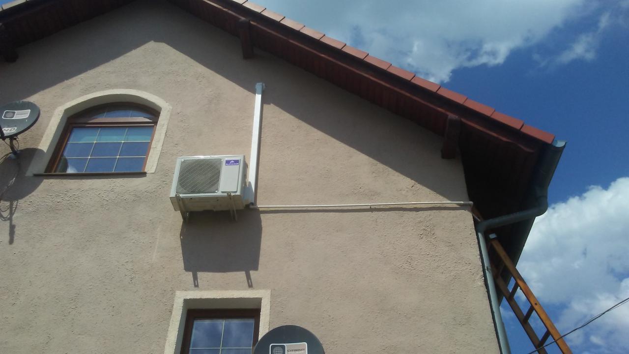 Zewnętrzna jednostka klimatyzacji zamontowana na elewacji domu z widocznymi przewodami, obok okna z łukiem i anteną satelitarną, na tle błękitnego nieba z chmurami.