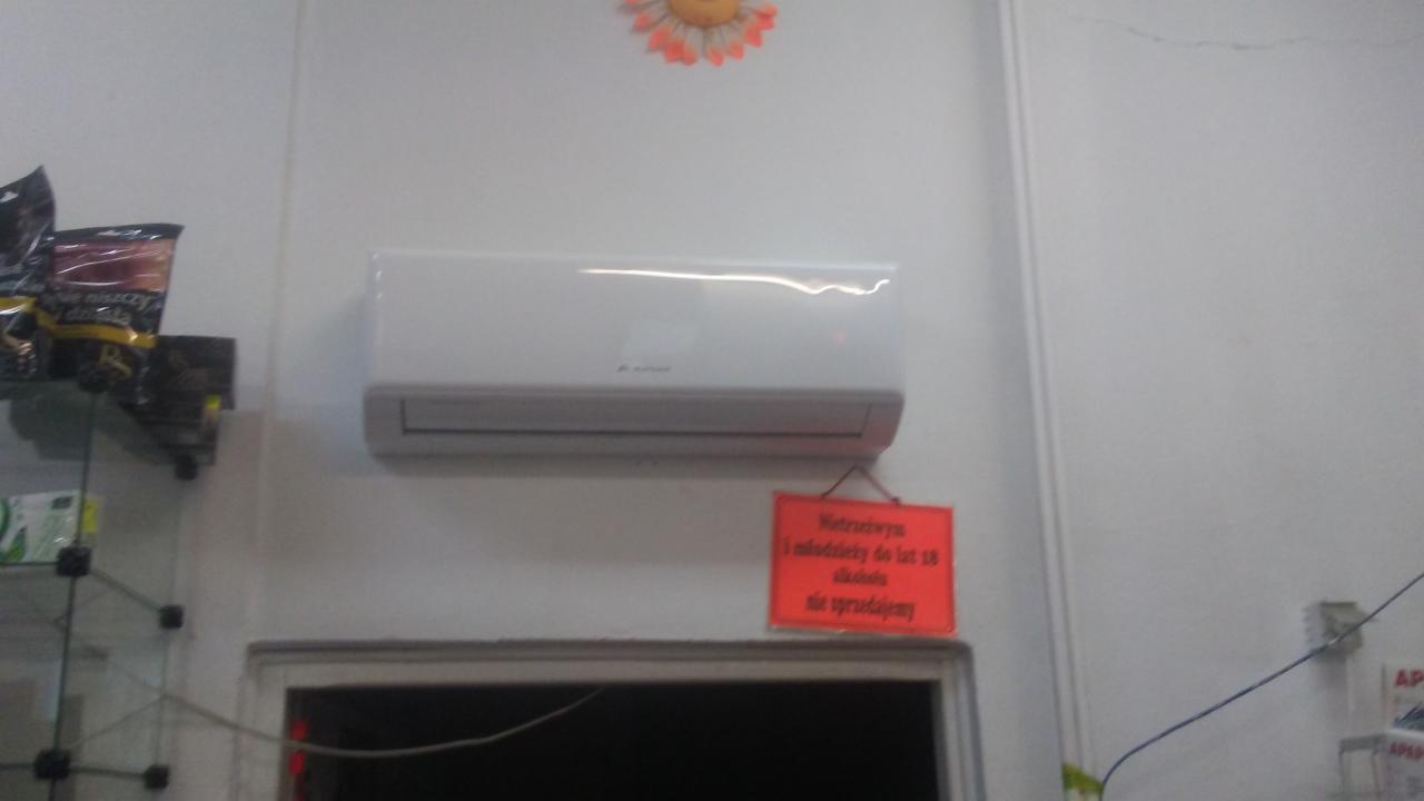 Biały klimatyzator ścienny zamontowany nad drzwiami w pomieszczeniu z półkami, na ścianie wisi czerwona tabliczka informacyjna.