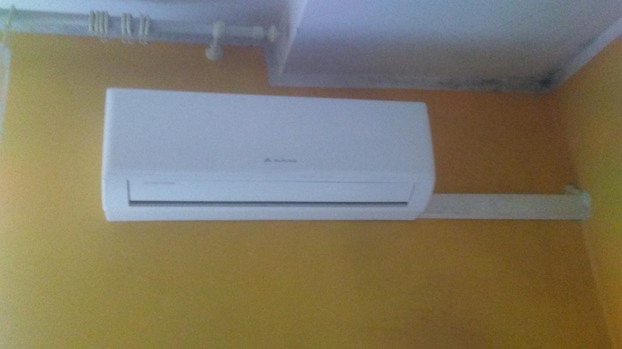 Zamontowany biały klimatyzator na żółtej ścianie w pomieszczeniu, widoczne rury pod sufitem i ślady wilgoci na suficie.