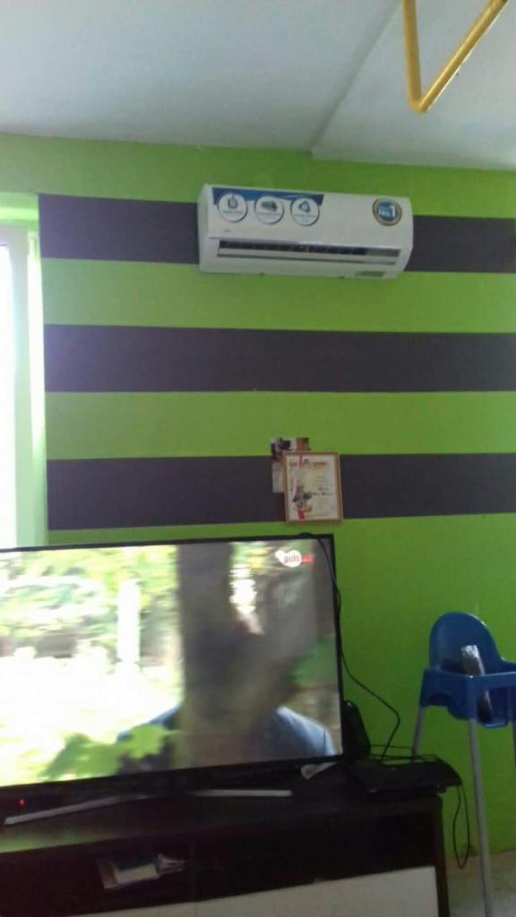 Klimatyzator ścienny zamontowany na zielono-szarej ścianie w pokoju, widoczny telewizor i krzesełko dziecięce.