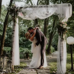 Pocałunek pary młodej pod drewnianym, udekorowanym kwiatami i białym tiulem łukiem na tle zieleni parku.