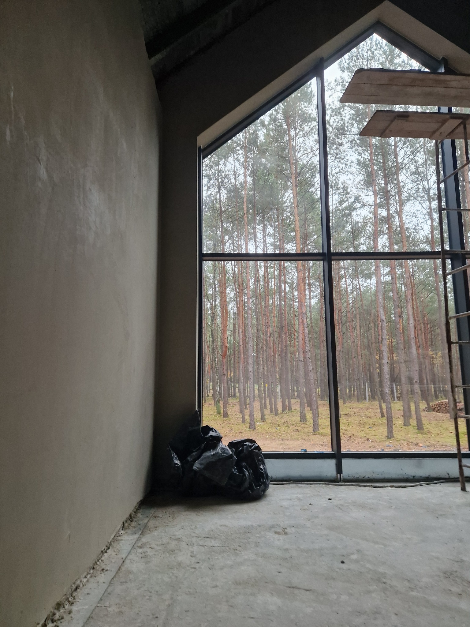 Świeżo otynkowana ściana w surowym wnętrzu z dużym oknem, przez które widać las; worki z odpadami budowlanymi w rogu, fragment rusztowania przy oknie.