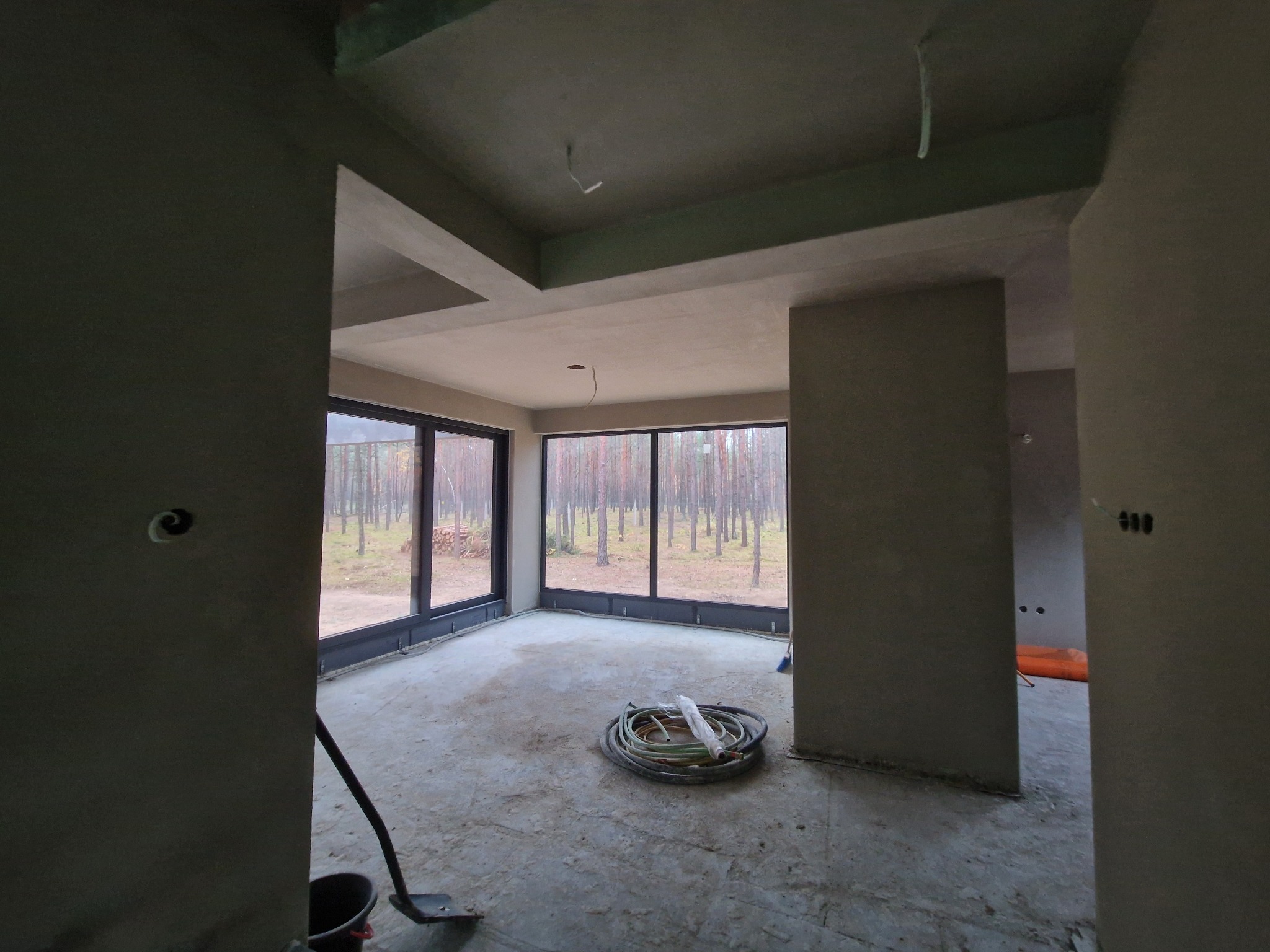 Surowe wnętrze w stanie deweloperskim z wygładzonymi ścianami i sufitem, widoczne przewody elektryczne, okna z widokiem na las, na podłodze zwinięty wąż i wiadro z narzędziami.