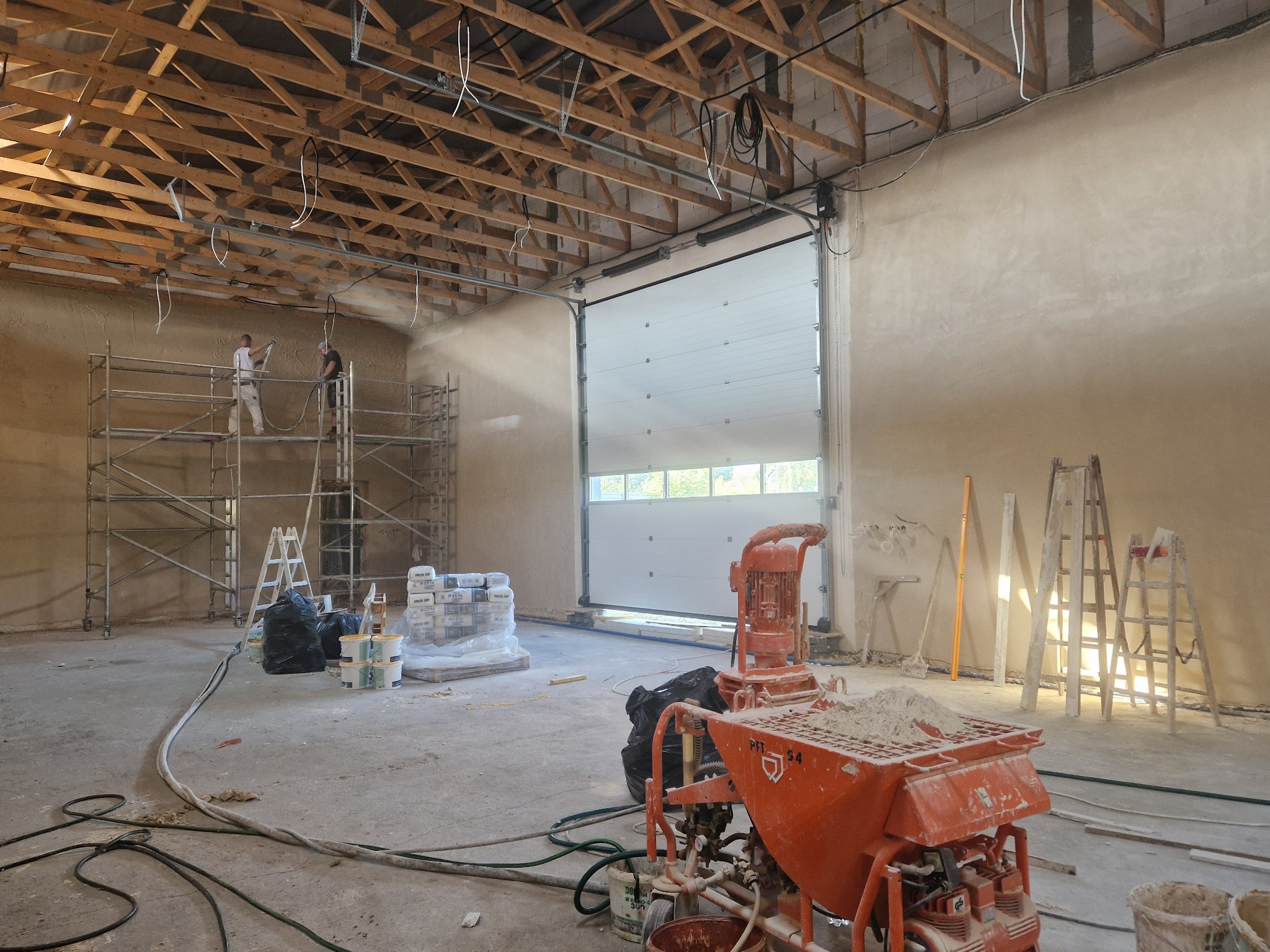 Wnętrze w trakcie prac wykończeniowych: dwóch pracowników na rusztowaniu aplikuje tynk na ścianę, w tle brama garażowa, na pierwszym planie maszyna do tynkowania i narzędzia.