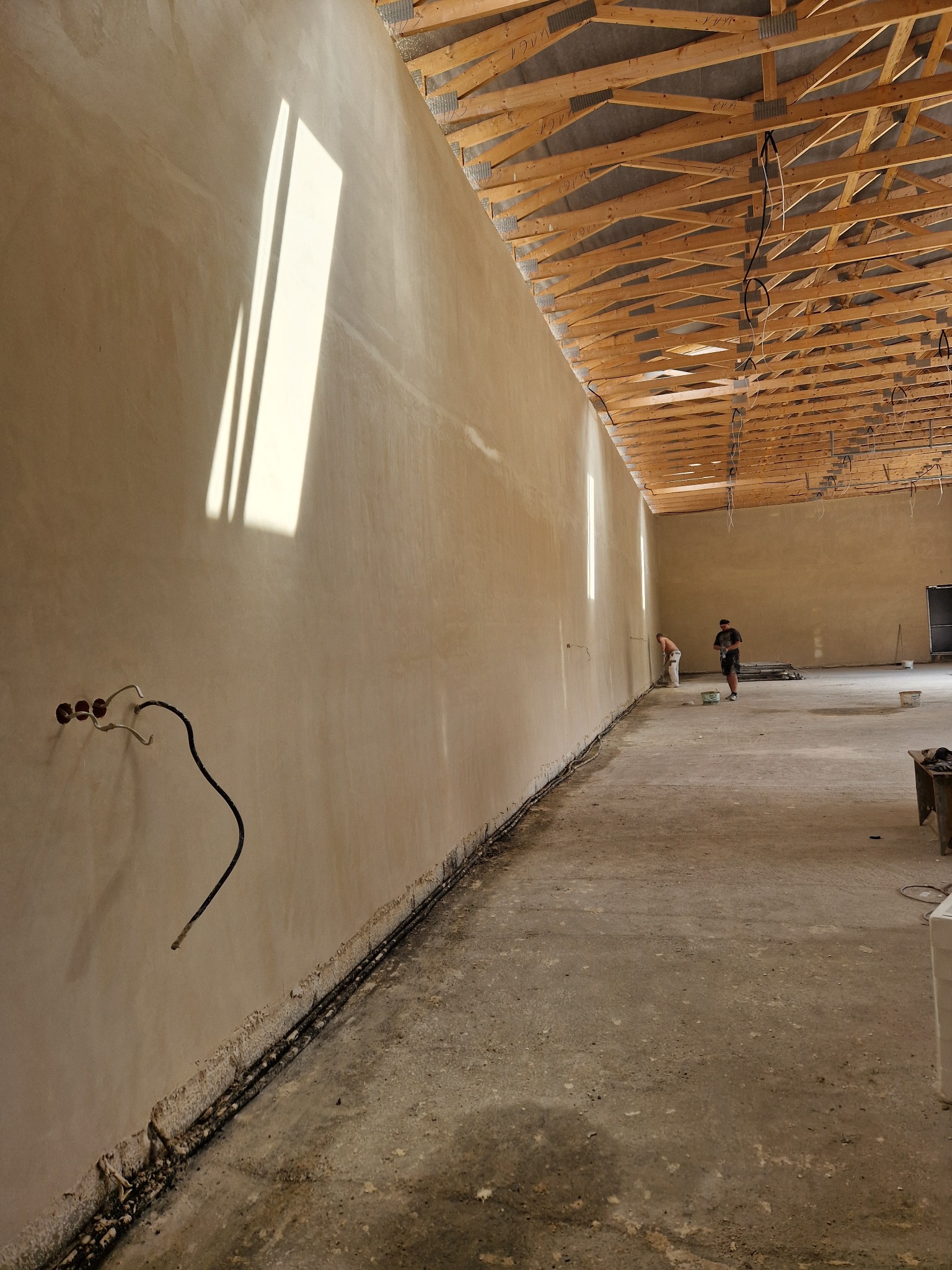 Wnętrze hali w trakcie tynkowania maszynowego; widoczne ślady po instalacjach elektrycznych, surowy beton na podłodze i drewniana konstrukcja dachu.