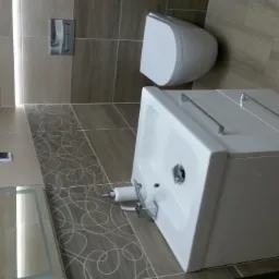 Nowoczesna łazienka z beżowymi płytkami imitującymi drewno, podwieszana toaleta i biała umywalka z szafką, na podłodze dekoracyjny wzór.