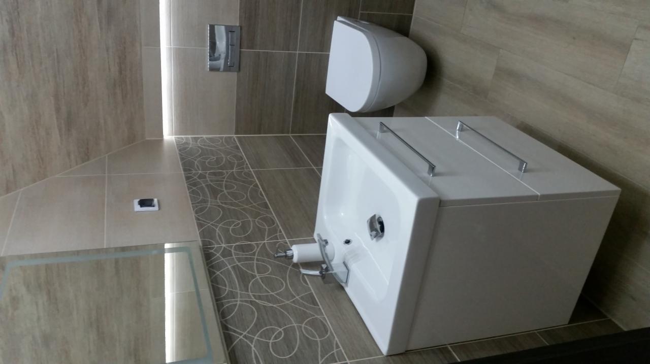 Nowoczesna łazienka z beżowymi płytkami imitującymi drewno, podwieszana toaleta i biała umywalka z szafką, na podłodze dekoracyjny wzór.