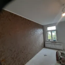 Pokój po remoncie w Ostrzeszowie z dekoracyjną ścianą w odcieniach brązu z motywem liści, widoczne worki z materiałami budowlanymi na podłodze i grzejnik pod oknem.
