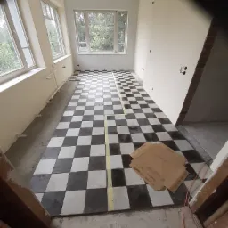 Pok&oacute;j z nowo ułożoną, czarno-białą szachownicą na podłodze, zabezpieczoną taśmą malarską, z widocznymi oknami i przygotowanymi ścianami do dalszych prac wykończeniowych.