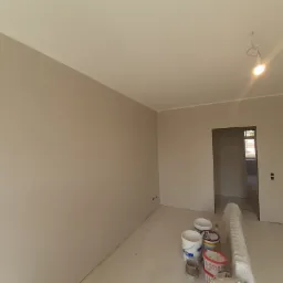 Wnętrze pokoju w trakcie odświeżania, ściany pomalowane na jasny beż, widoczne wiadra z farbą oraz grzejnik zabezpieczony folią.