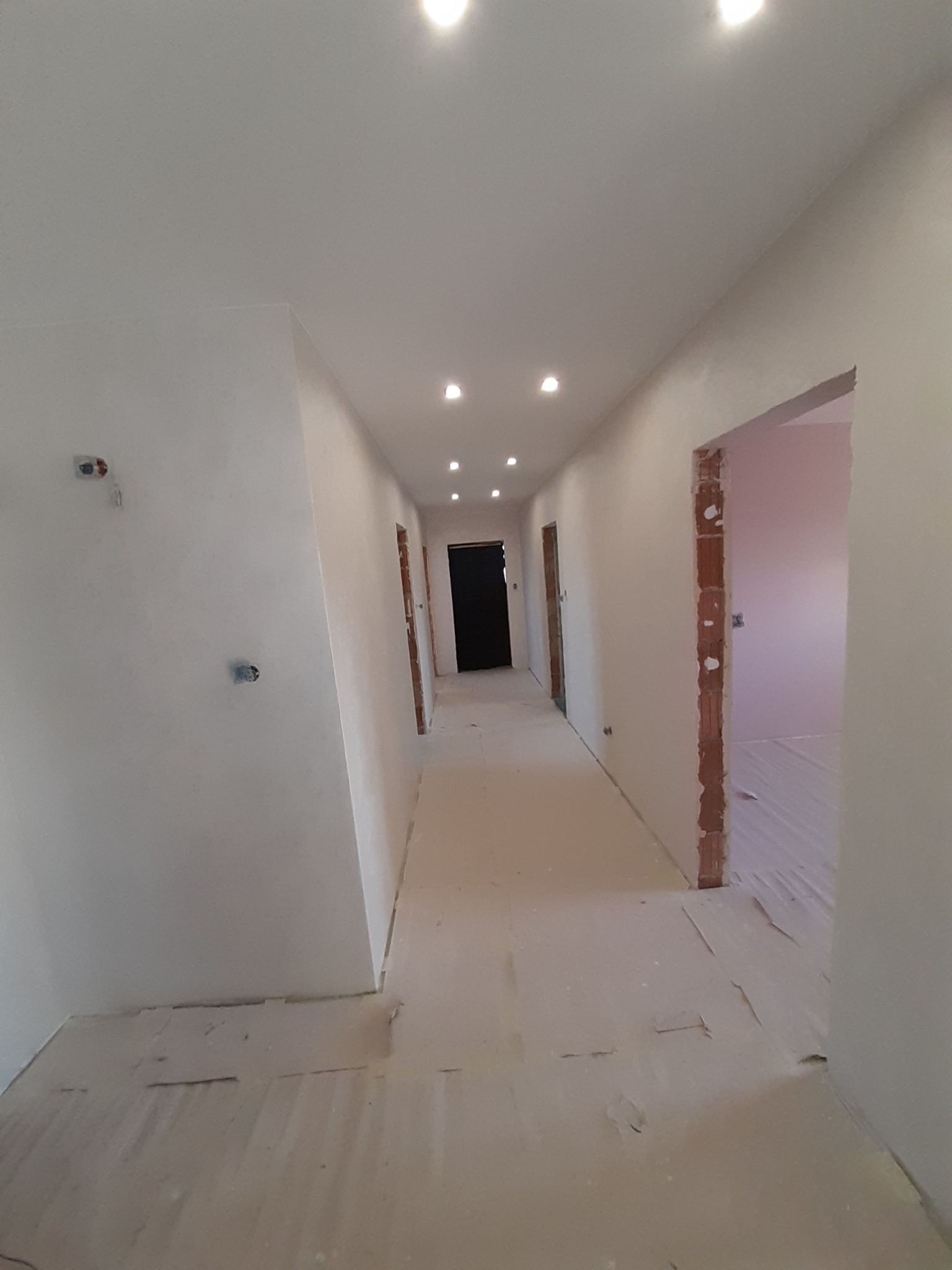 Korytarz w trakcie wykańczania: ściany po szpachlowaniu, widoczne puszki elektryczne, podłoga zabezpieczona folią malarską, oświetlenie sufitowe punktowe.
