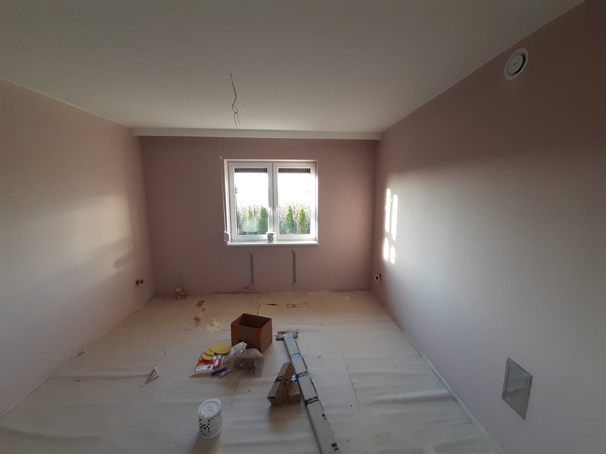 Pokój po malowaniu ścian na delikatny róż, zabezpieczony podłogą. Widoczne narzędzia malarskie, puszka z farbą i kartonowe pudło.