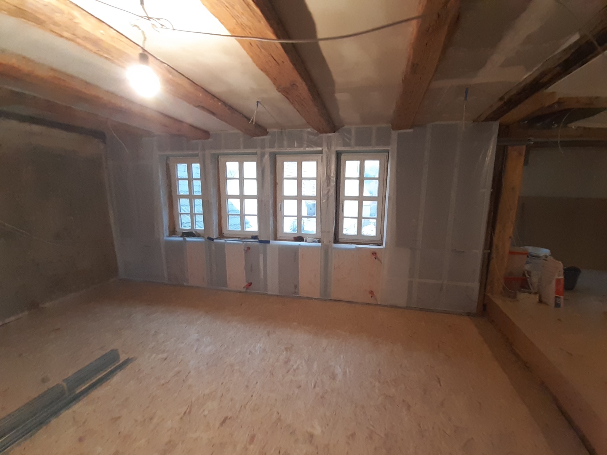 Wnętrze w trakcie prac wykończeniowych: ściany z płyt kartonowo-gipsowych, częściowo pokryte szpachlą, drewniane belki stropowe, trzy okna z widokiem na zewnątrz, podłoga z płyt OSB, widoczne...