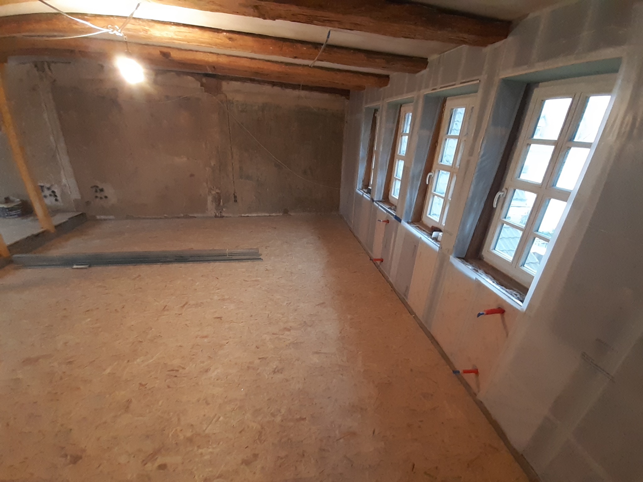 Wnętrze remontowanego poddasza z odsłoniętymi belkami stropowymi, płytami karton gips na ścianach przy oknach i surową podłogą z płyty OSB.