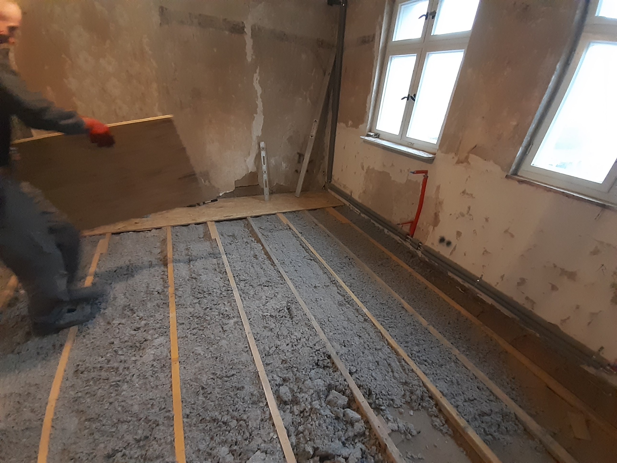 Pracownik w czerwonych rękawicach układa płytę OSB na izolacji z wełny mineralnej między legarami na podłodze w remontowanym pomieszczeniu z oknami i nieotynkowanymi ścianami.