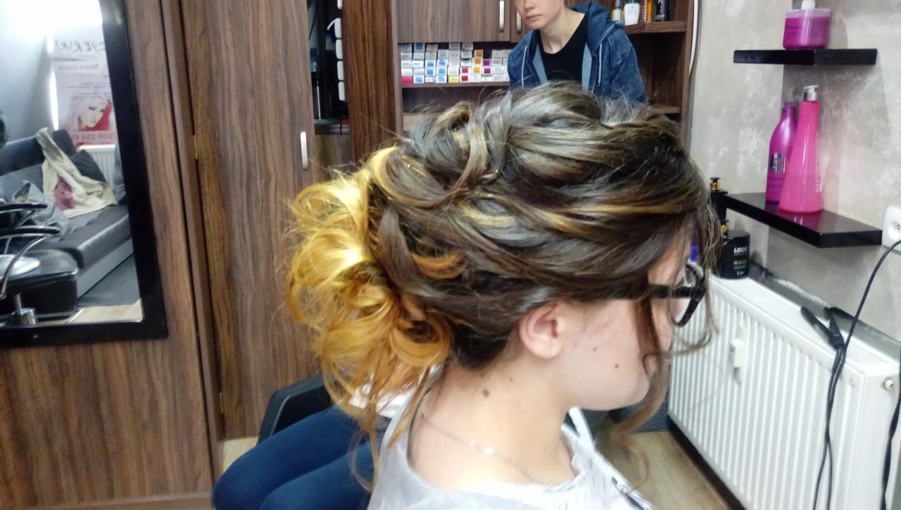 Upięcie włosów z pasemkami w odcieniach blondu, widok z boku na klientkę w okularach w salonie, w tle regał z kosmetykami.