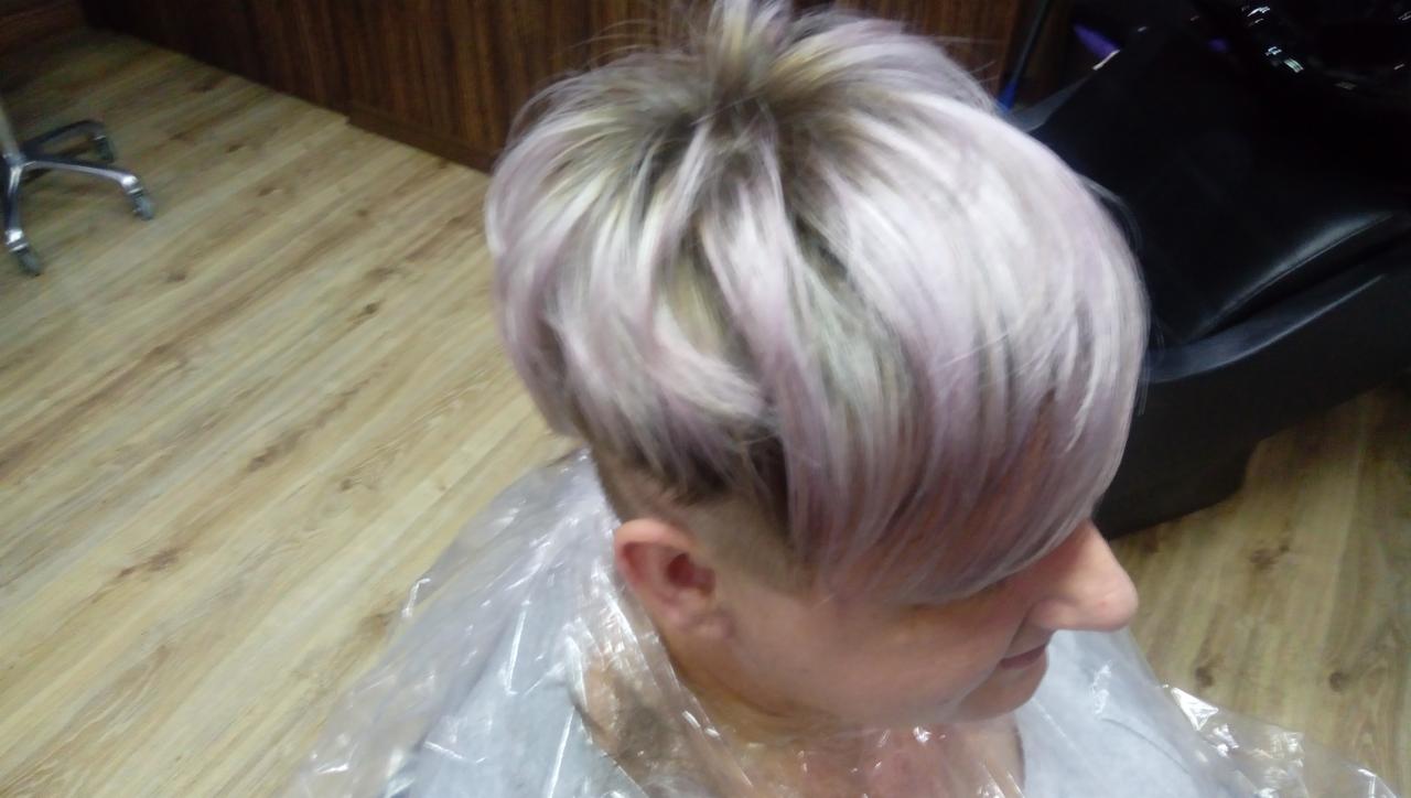Krótkie, teksturowane włosy w odcieniach platyny i fioletu, z widocznym podgoleniem na karku, klientka w pelerynie ochronnej podczas zabiegu.