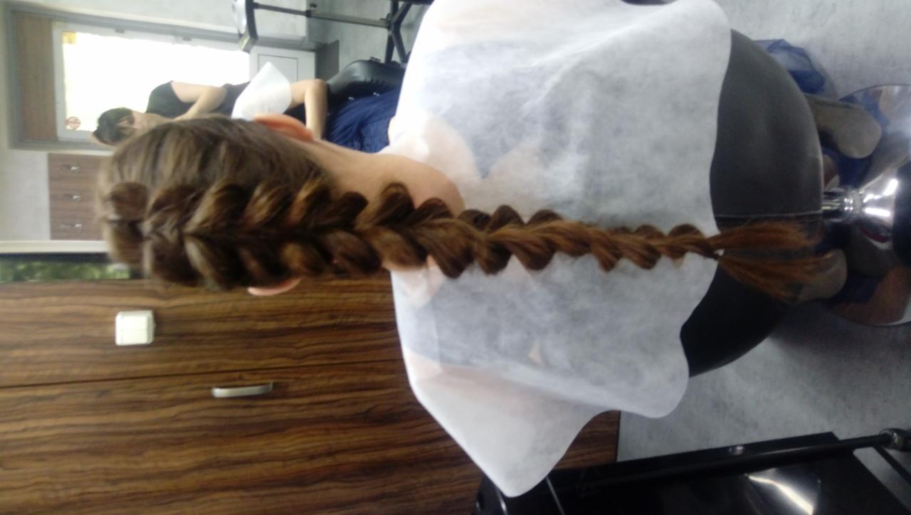 Długi, ciemny warkocz typu kłos, zapleciony na włosach klientki siedzącej w fotelu w salonie, widok z tyłu, z widoczną jednorazową peleryną ochronną.