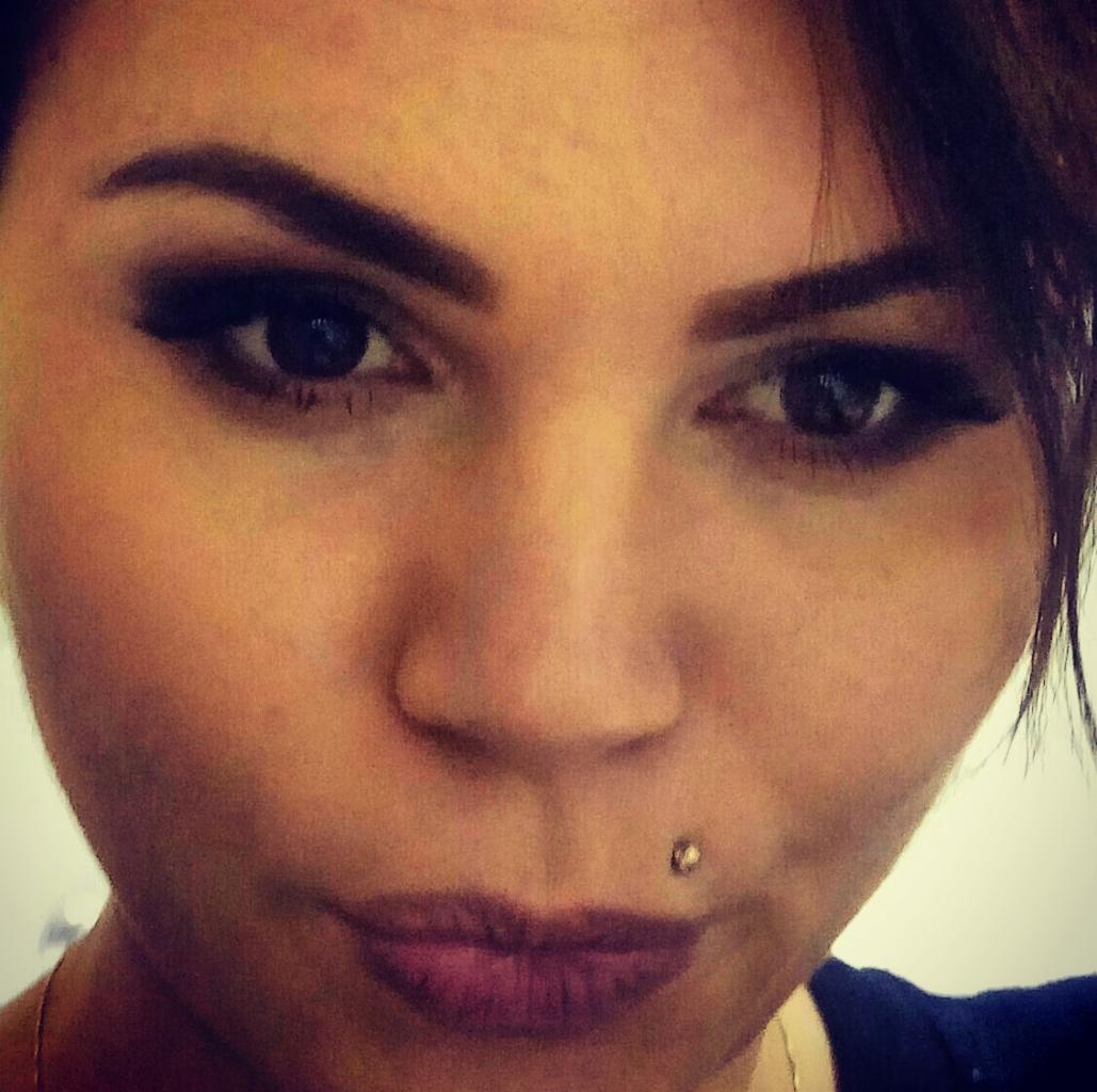 Portret kobiety z profesjonalnym makijażem oczu i ust, z kolczykiem w wardze. Widoczne perfekcyjnie wystylizowane brwi i delikatny cień do powiek.