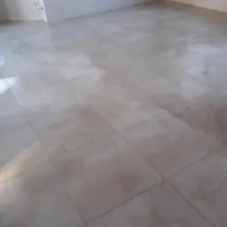 Jasne, błyszczące płytki podłogowe imitujące marmur, ułożone w kwadraty z ciemniejszymi fugami, wypełniające puste pomieszczenie z fragmentem białej ściany u góry i krawędzią białej ściany po lewej...