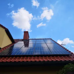 Panel słoneczny na czerwonym dachu domu jednorodzinnego, widok z dołu, błękitne niebo z chmurami w tle.