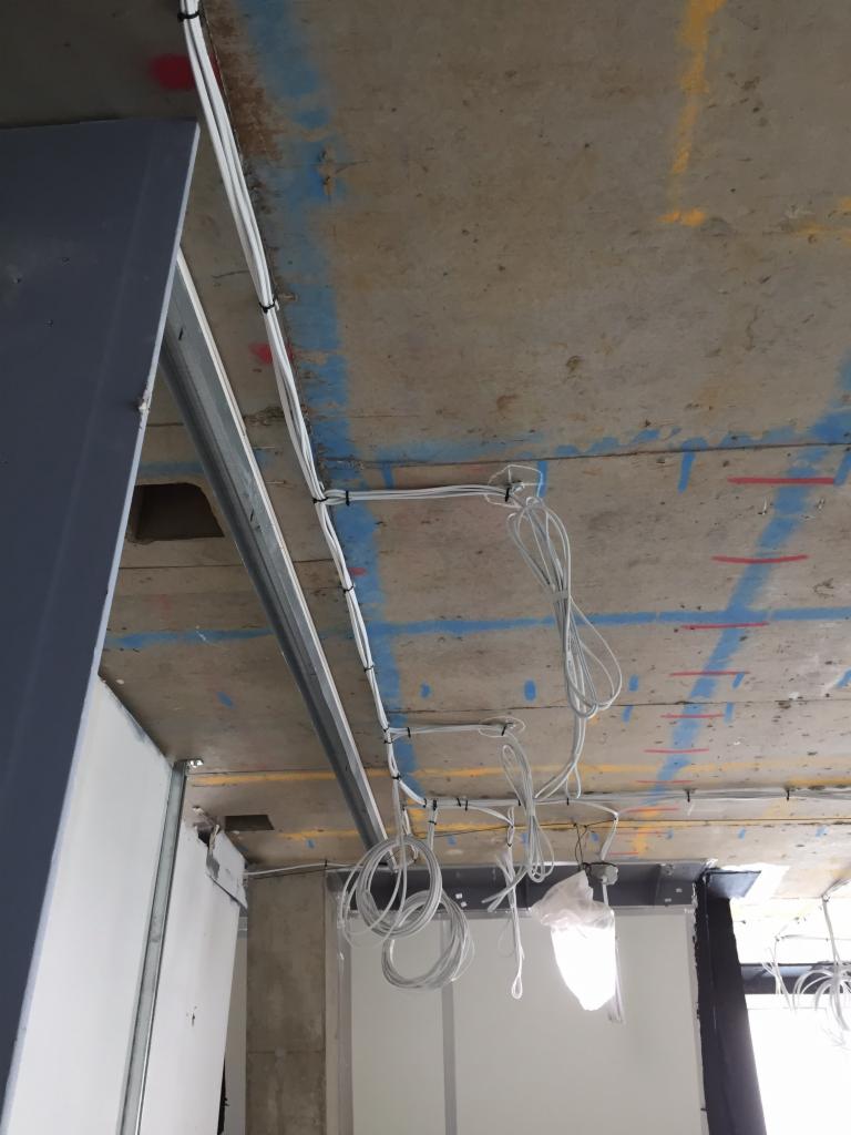Surowy betonowy sufit z widocznymi przewodami elektrycznymi mocowanymi do konstrukcji metalowej, w tle fragment ściany z płyt gipsowo-kartonowych.