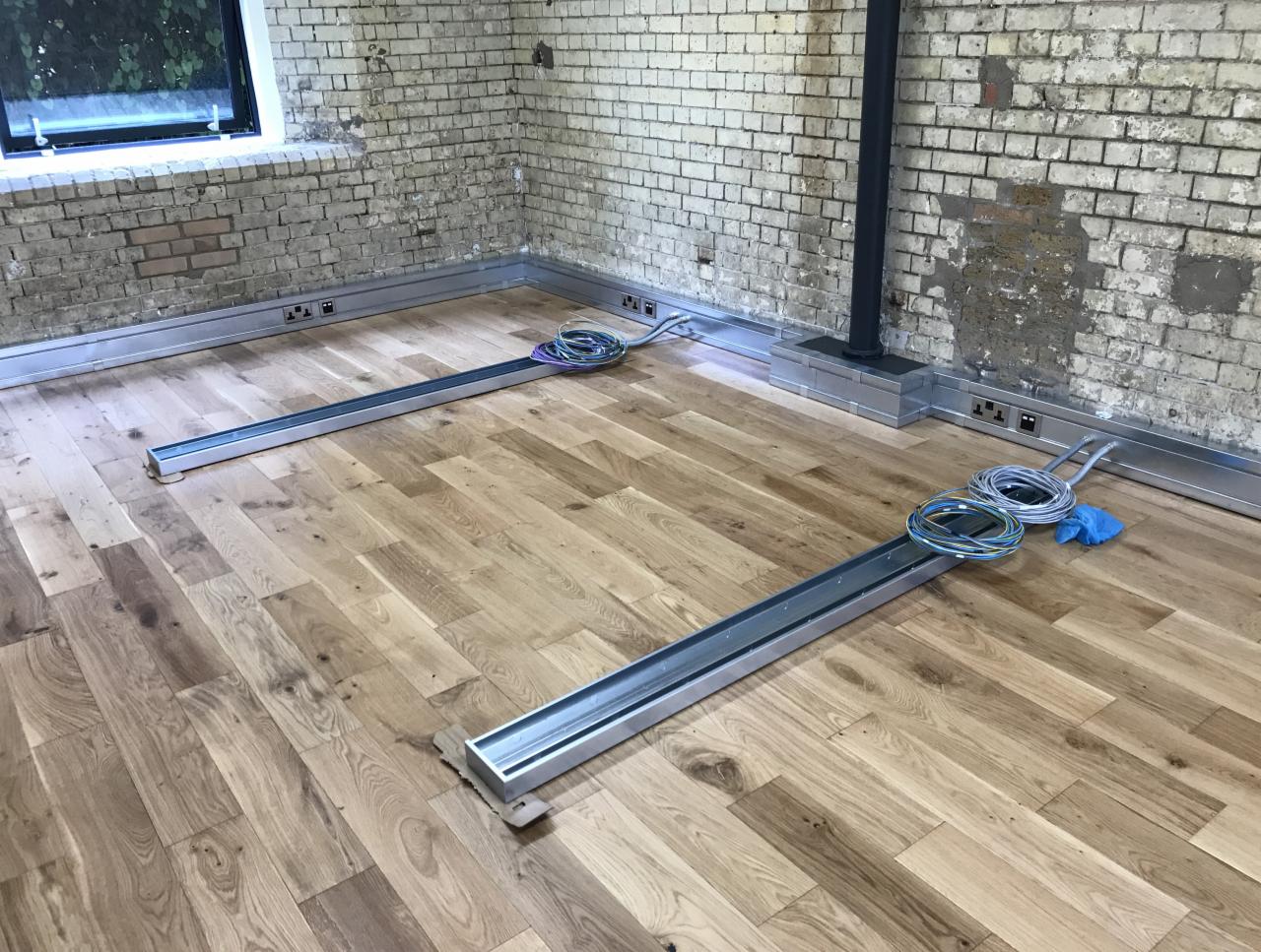 Fragment pomieszczenia z drewnianą podłogą i surową ceglaną ścianą, widoczne aluminiowe korytka pod kable elektryczne w trakcie montażu, zwinięte przewody i niebieska ściereczka leżą na podłodze.