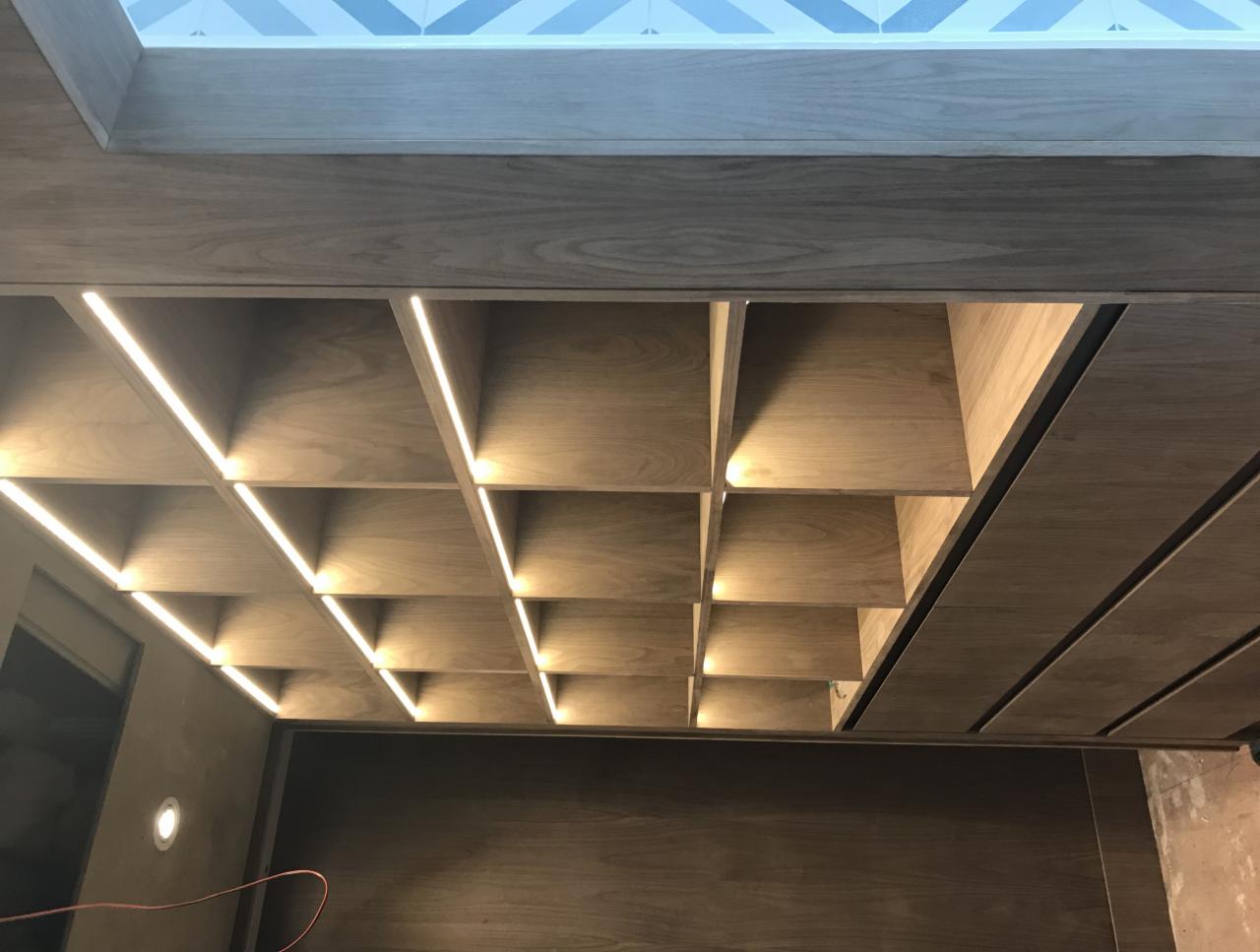 Drewniany sufit kasetonowy z oświetleniem LED w szczelinach, widok z dołu w trakcie montażu, widoczny fragment sufitu z niebiesko-białą mozaiką.