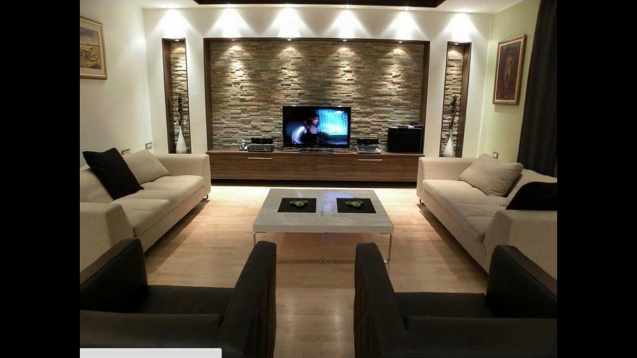 Nowoczesny salon z kamienną ścianą podświetloną punktowymi lampami, drewnianą szafką RTV, dwoma beżowymi sofami i ciemnymi fotelami, całość oświetlona ciepłym światłem.