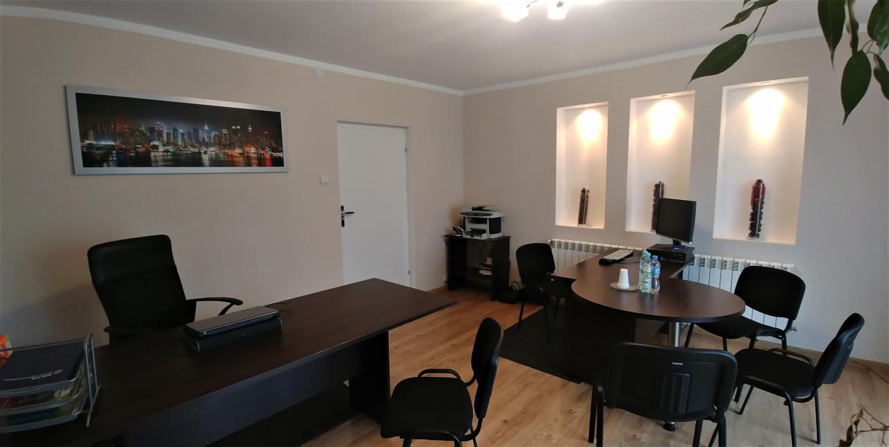 Biuro agencji nieruchomości z ciemnymi meblami, trzema wnękami z oświetleniem i dekoracjami, obrazem panoramy miasta na ścianie oraz rośliną doniczkową w rogu pomieszczenia.