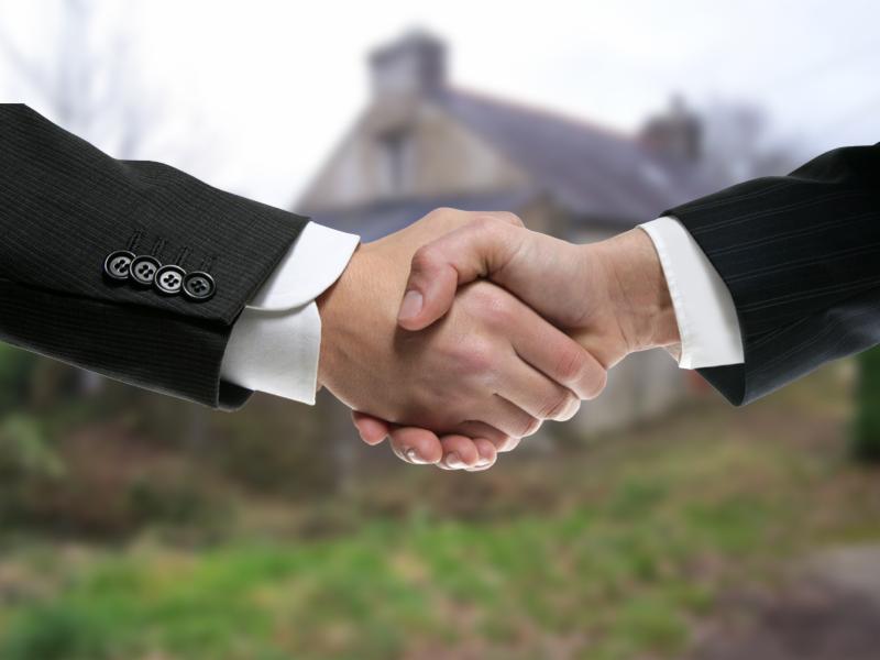 Uścisk dłoni na tle rozmytego domu z dwoma kominami, widoczny mankiet koszuli i marynarki w paski.