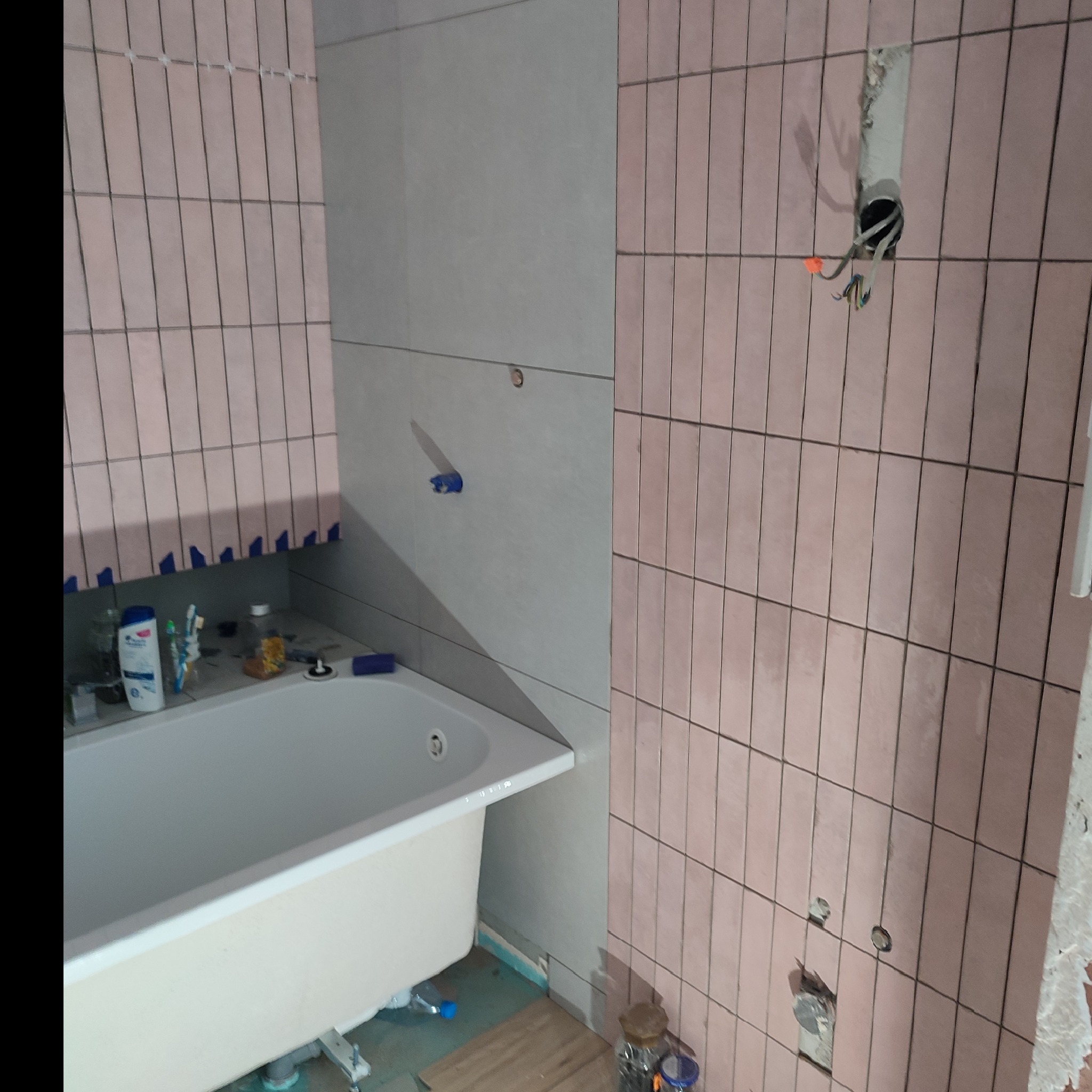 Łazienka w trakcie remontu: biała wanna z kosmetykami na krawędzi, różowe pionowe płytki częściowo ułożone, widoczne przewody elektryczne i otwory pod armaturę.