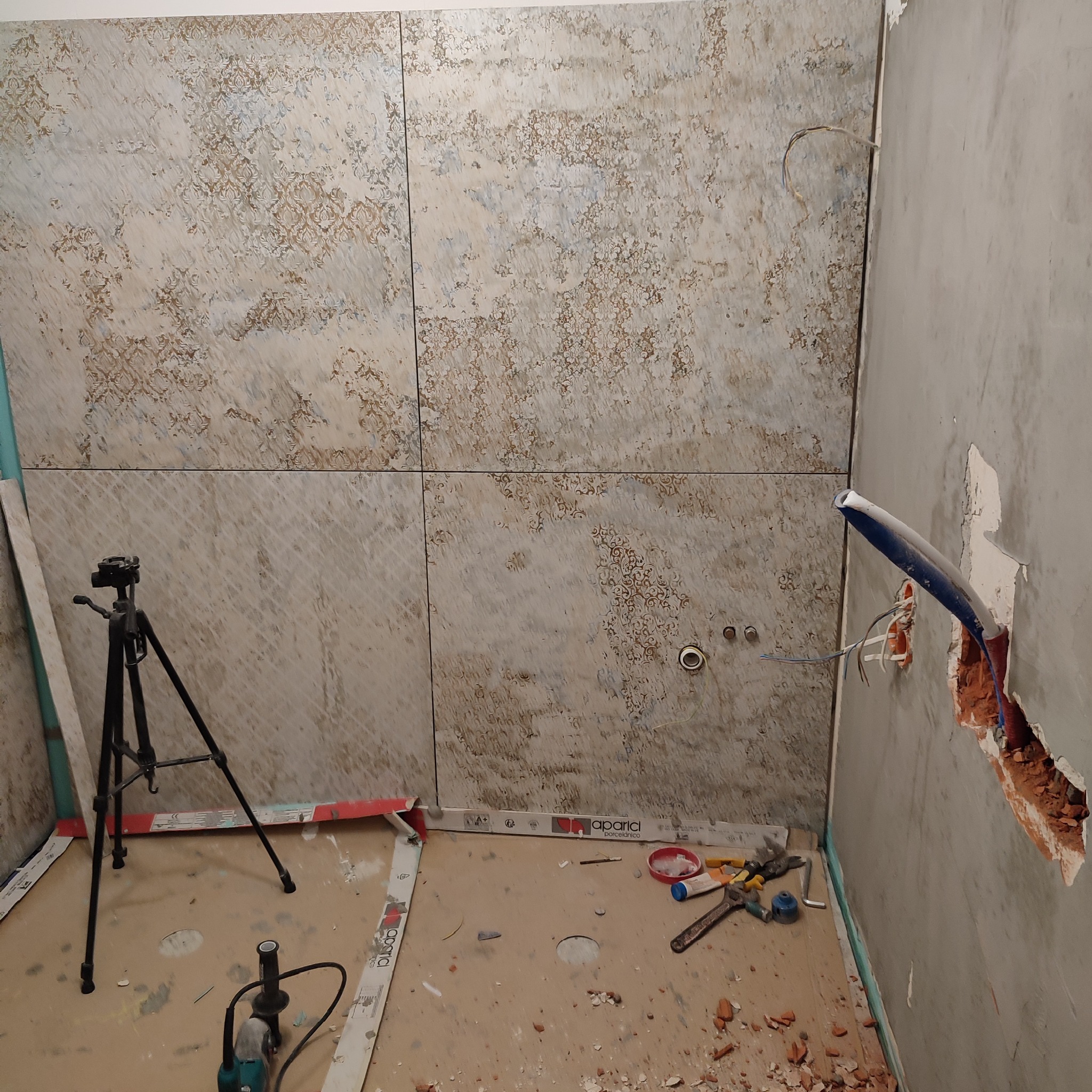 Ściana łazienki w trakcie wykańczania z położonymi płytkami o wzorzystym deseniu, widoczne wystające rury, narzędzia i statyw na podłodze.