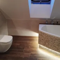 Wykończona łazienka na poddaszu z drewnopodobną podłogą, mozaiką na obudowie wanny z podświetleniem LED i podwieszanym WC.