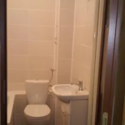 Widok na małą, wykończoną łazienkę z beżowymi płytkami, białym wyposażeniem i drewnopodobną podłogą, widzianą zza futryny drzwi.