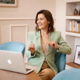 Uśmiechnięta kobieta w pastelowej marynarce siedzi przy biurku z laptopem, trzymając okulary w dłoniach i spoglądając w bok.