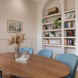 Eleganckie wnętrze: owalny stół z drewna orzechowego, błękitne krzesła, wazon z suszonymi trawami, w tle regał z książkami i staromodne radio.